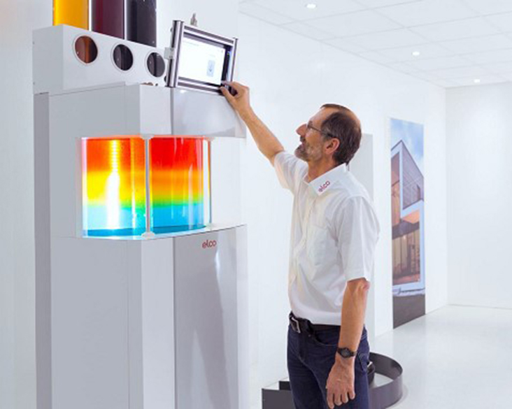 Person demonstriert ein ELCO Heizsystem mit digitalem Bedienfeld und visualisierter Wärmeverteilung in einem Ausstellungsraum.